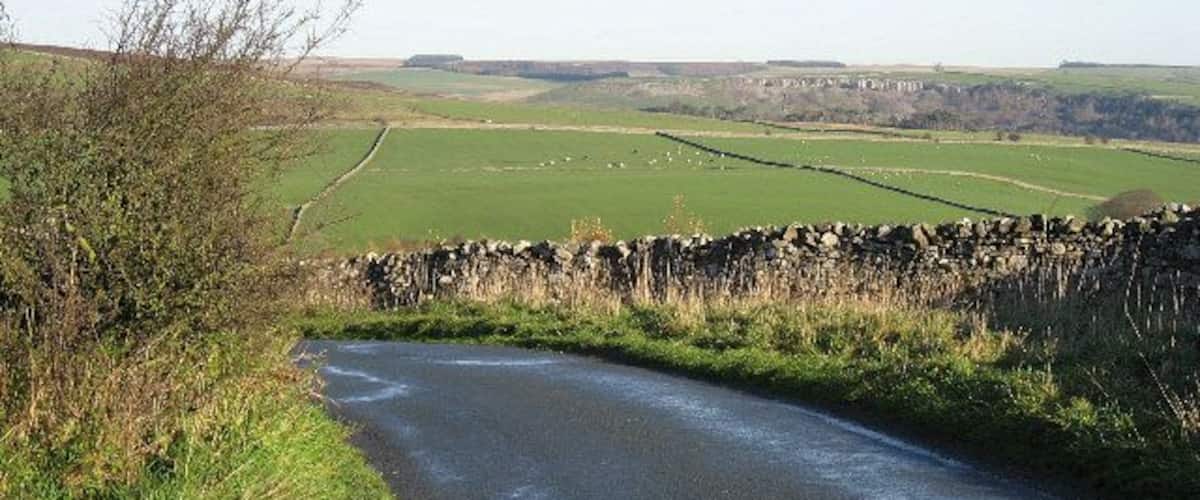 A narrow rural road curving through green fields with stone walls and a stone bridge.