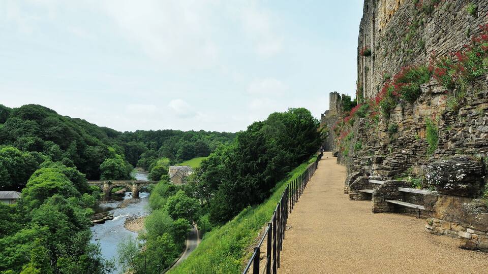 A stone pathway alongside a river, a bridge, and a castle wall with vegetation.