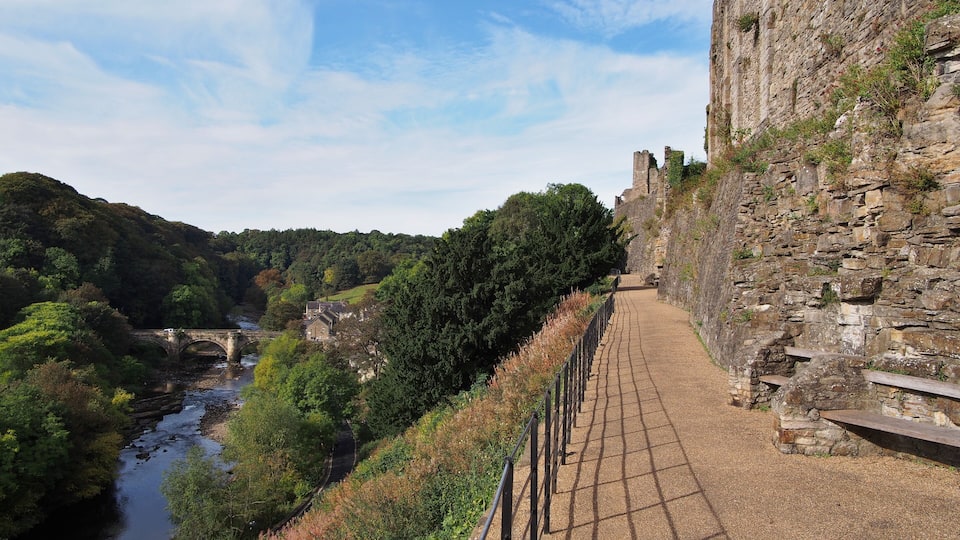 A stone pathway beside a river, leading to a historic stone wall and a bridge.