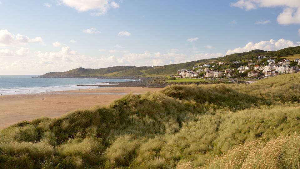 A coastal landscape with a sandy beach, grassy dunes, and a cluster of houses on a hillside.