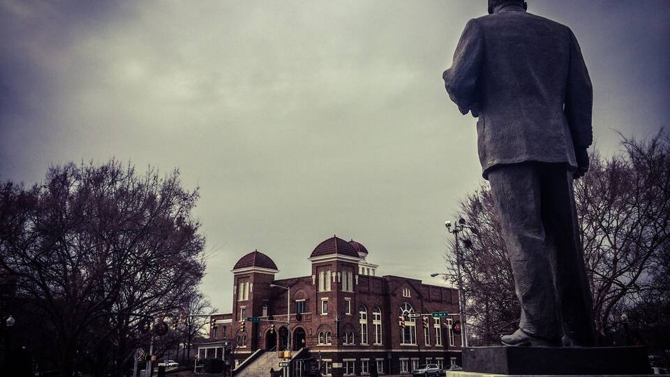 A statue of a man in a suit standing in front of a large brick building with arched windows and domed roofs.