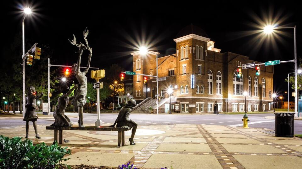A nighttime cityscape with a historic building, streetlights, traffic lights, and a statue of a seated figure.