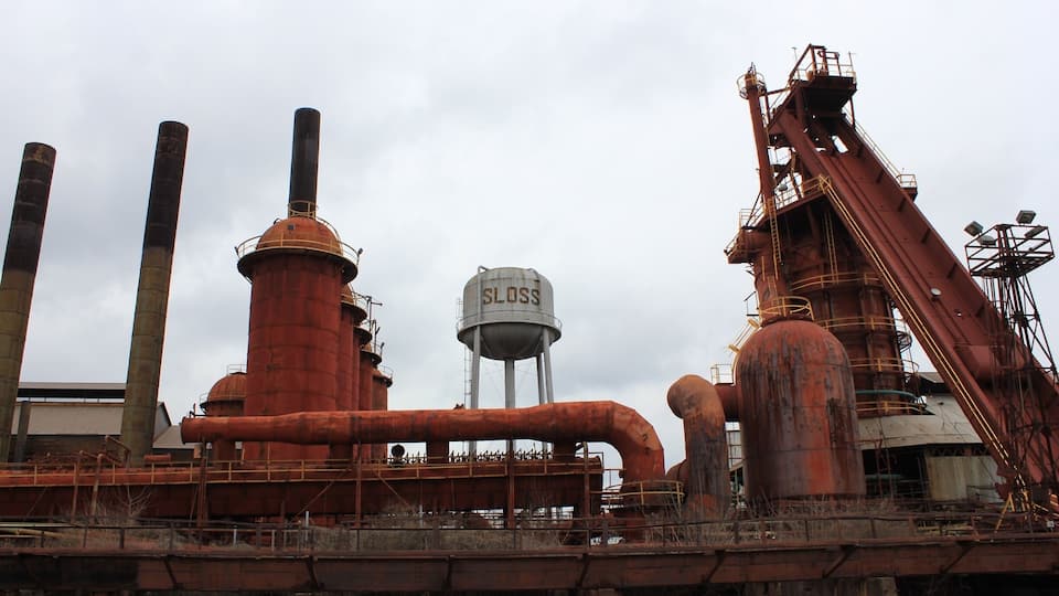 An industrial complex with rusted metal structures, including large pipes and a water tower labeled "SLOSS".