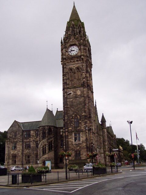 A historic clock tower with a pointed roof and a large clock face, surrounded by a building with multiple windows and a green roof.