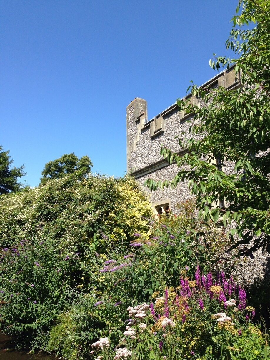 A historic stone building with a tower, surrounded by lush greenery and flowering plants.