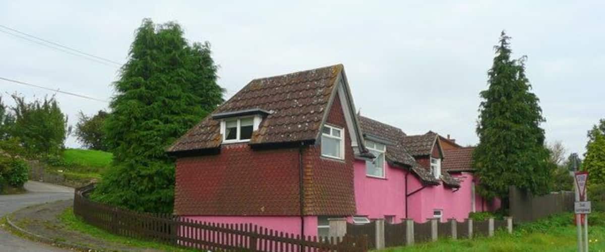 A pink house with a brown roof, surrounded by a fence and trees.