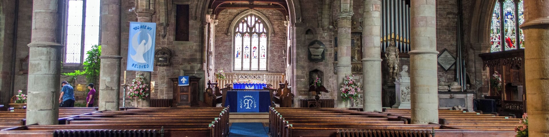 An interior of a church with arched ceilings, wooden pews, stained glass windows, and a central aisle leading to an altar.