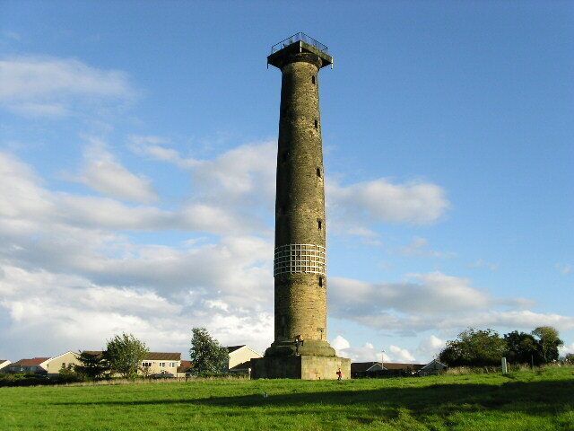 A tall, cylindrical stone tower with a metal railing at the top, surrounded by a grassy field and houses in the background.