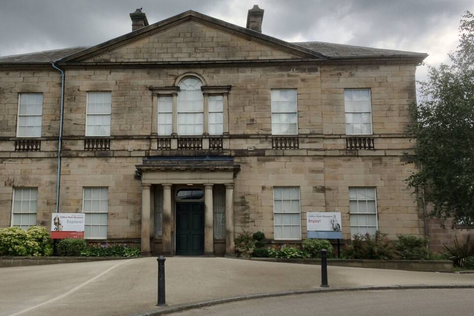 A historic stone building with a central entrance, flanked by two signs advertising the Clifton Park Museum.