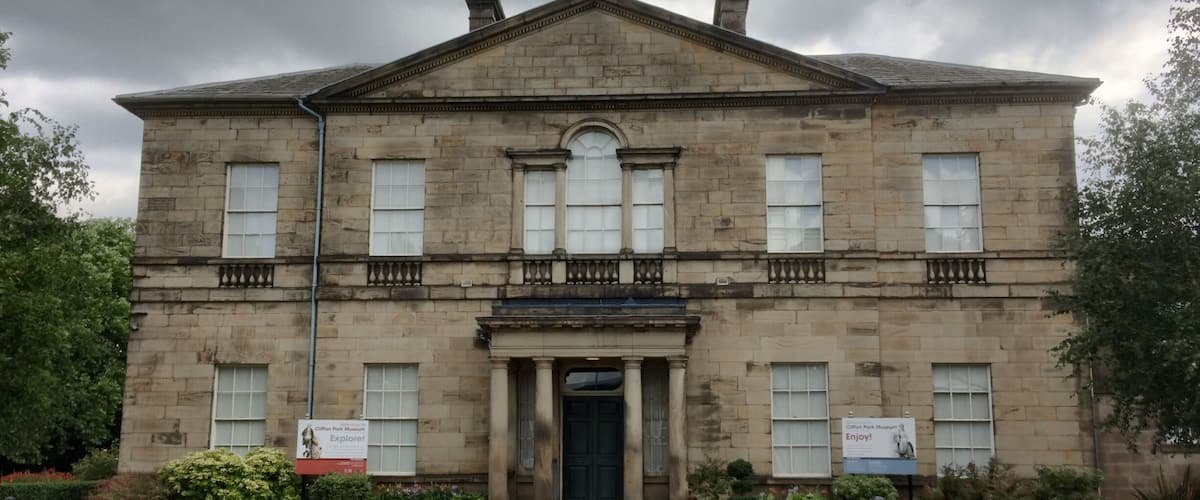 A historic stone building with a central entrance, flanked by two signs advertising the Clifton Park Museum.
