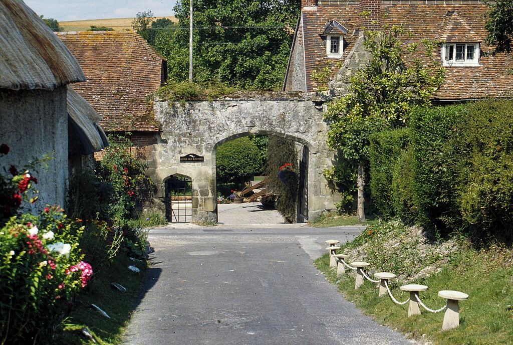 A stone archway leading to a village with traditional houses and a pathway lined with flowers.