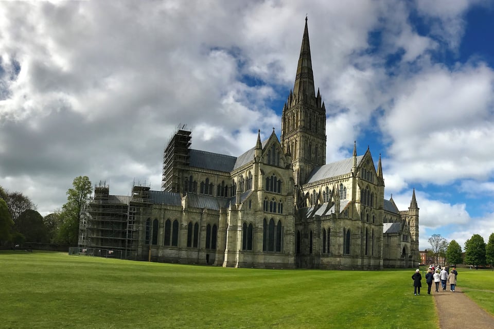Une cathédrale historique à l’architecture gothique, avec ses arcs en ogive et son flèche imposante, entourée d’une pelouse verdoyante et se dressant sous un ciel partiellement nuageux.