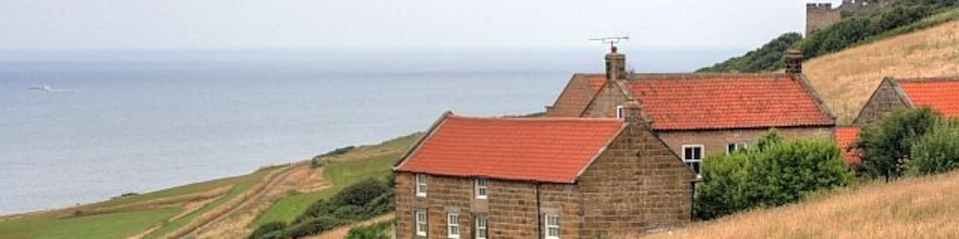 A stone house with a red roof on a grassy hill overlooking the sea.