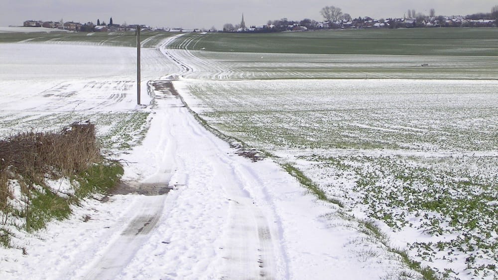 Eine verschneite Landstraße mit Feldern an beiden Seiten und einem Dorf in der Ferne.