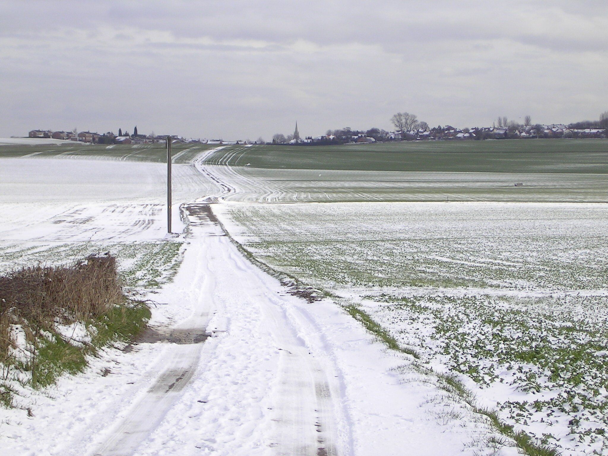 A snow-covered rural road with fields on either side and a distant village.