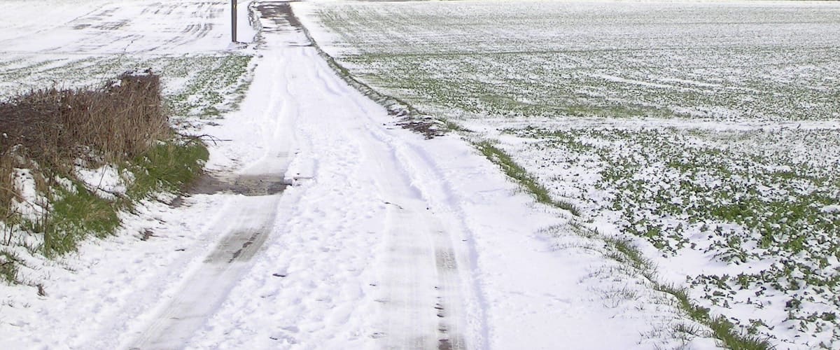 A snow-covered rural road with fields on either side and a distant village.