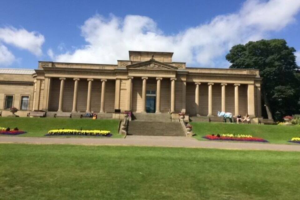 A neoclassical building with a central staircase, flanked by symmetrical columns, and a well-maintained lawn with flower beds.