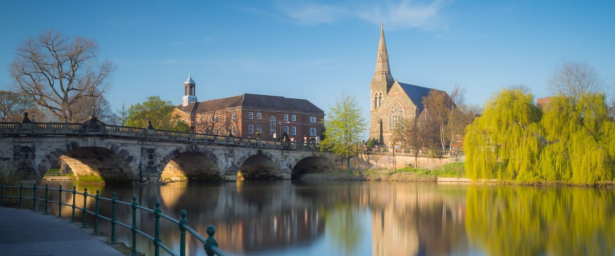 View along the river Severn to the English Bridge, Shrewsbury and a church and prominent willow tree, England, UK.