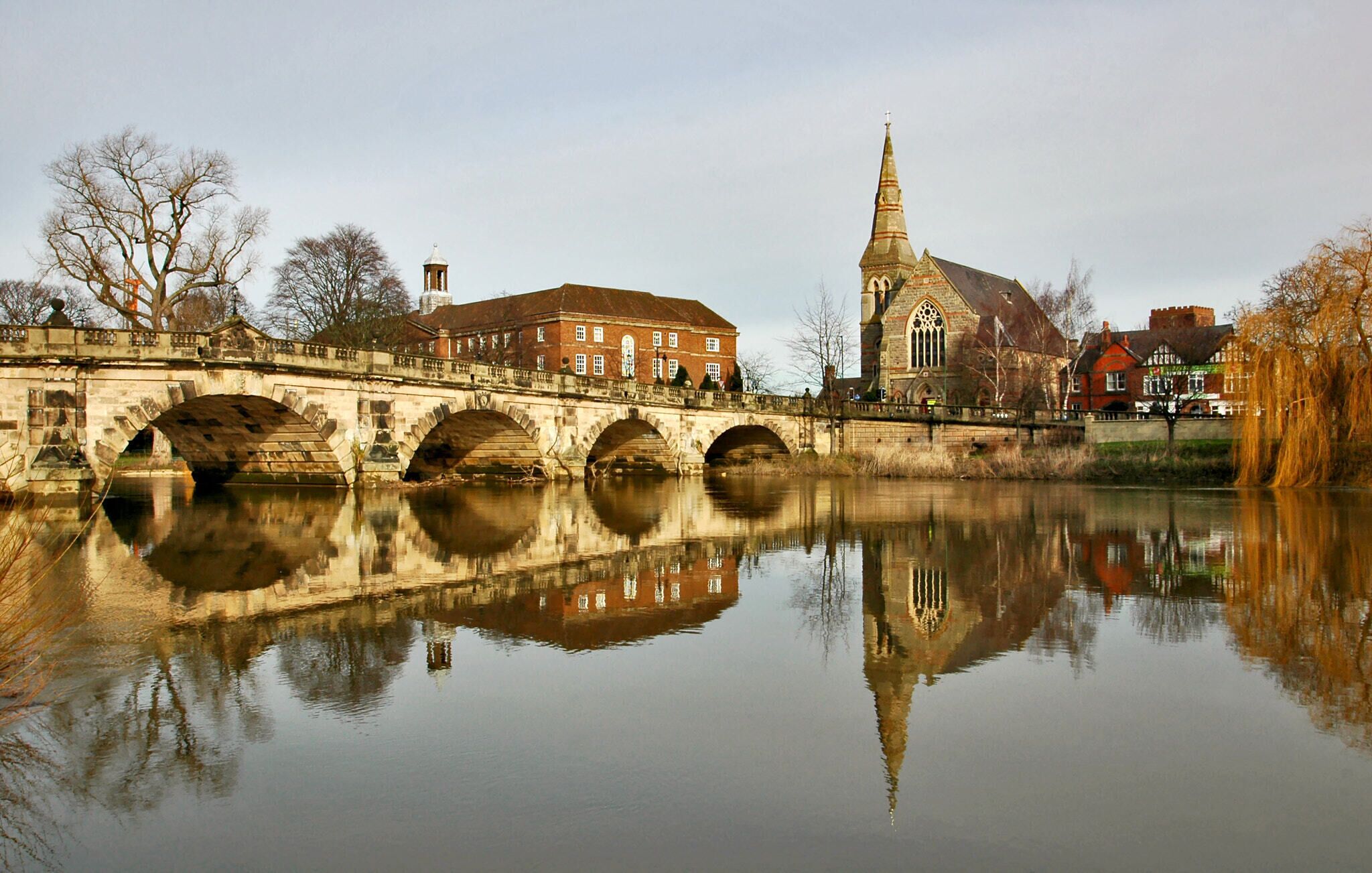 A stone bridge over a calm river, reflecting buildings and a church with a tall spire.