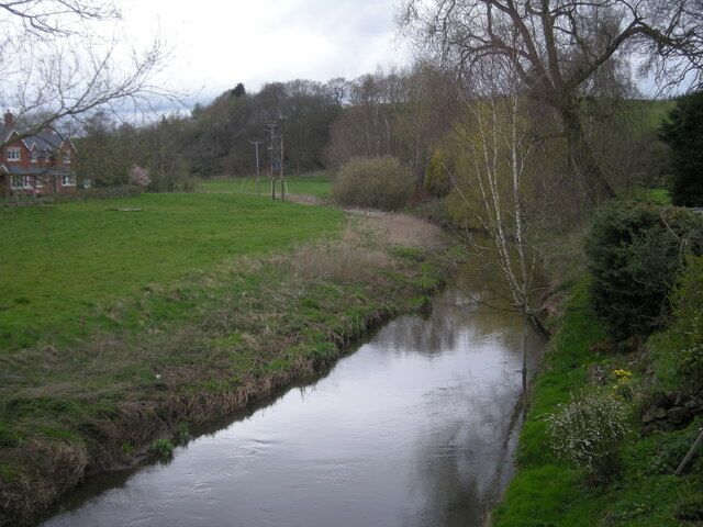 A narrow water channel with grassy banks, a red house in the background, and leafless trees.