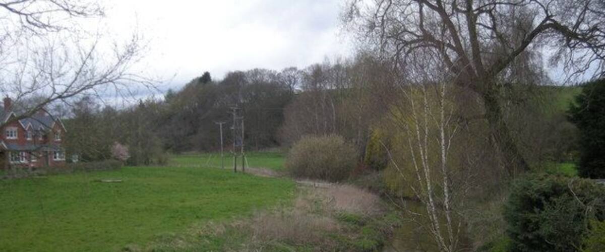 A narrow water channel with grassy banks, a red house in the background, and leafless trees.