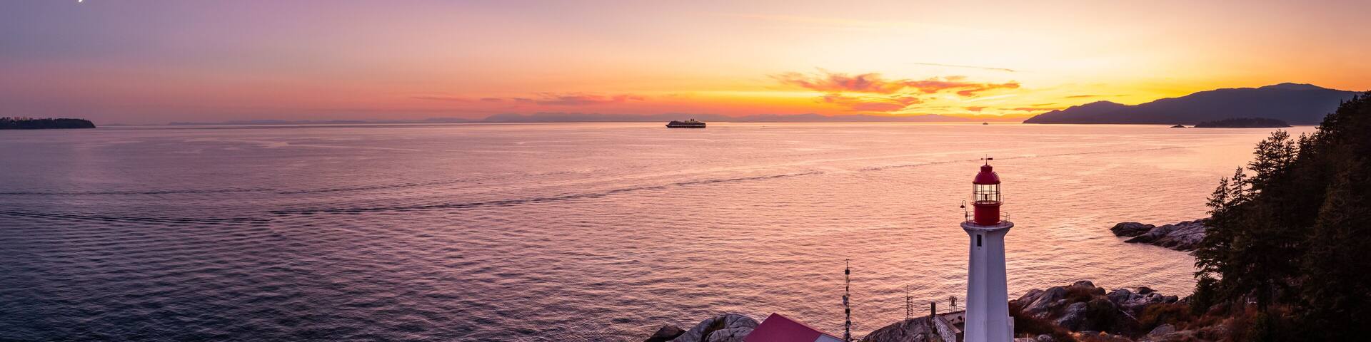 A lighthouse on a rocky coastline at sunset with calm waters and a distant ship.