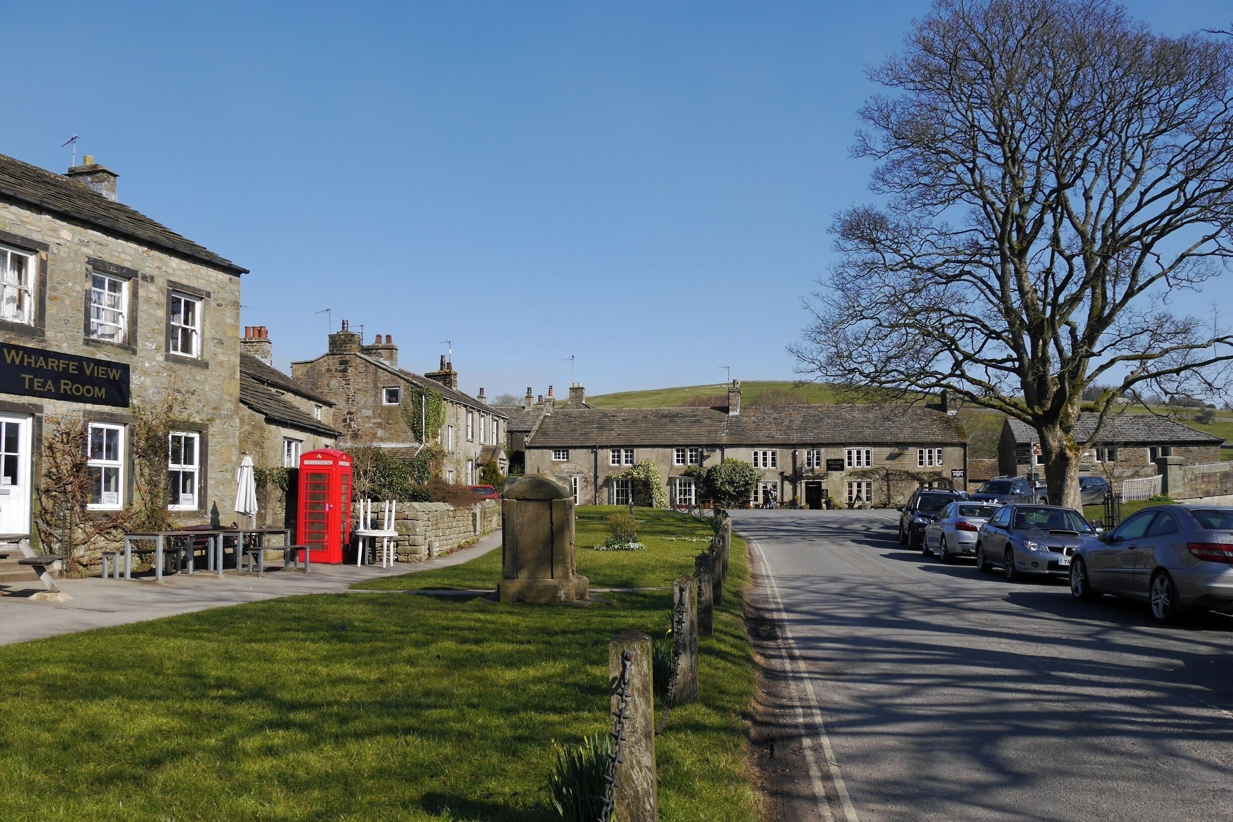 A quaint village street with stone buildings, a red telephone booth, and parked cars.