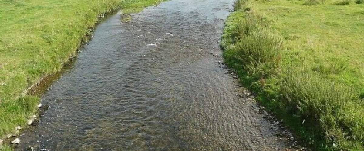 A narrow, clear stream flowing through a grassy field with a small building in the distance.