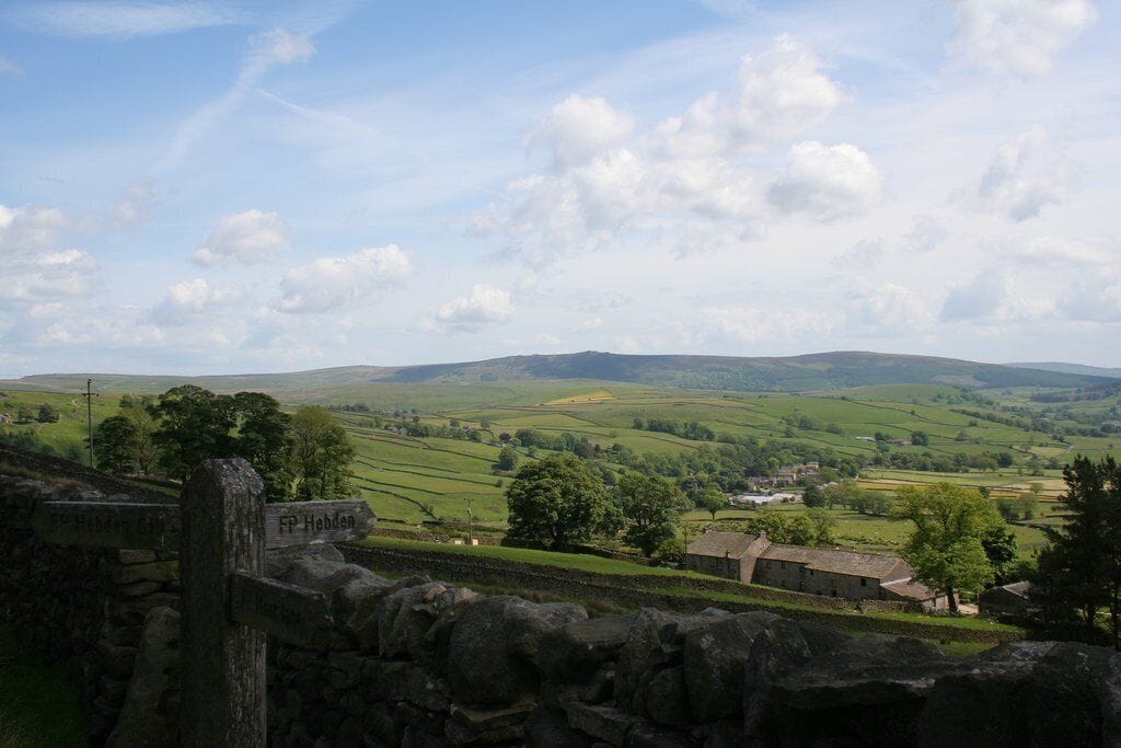 A stone wall with signs reading "FP Hobden" and "FP Hobden Gate" overlooking rolling green hills and a small village.