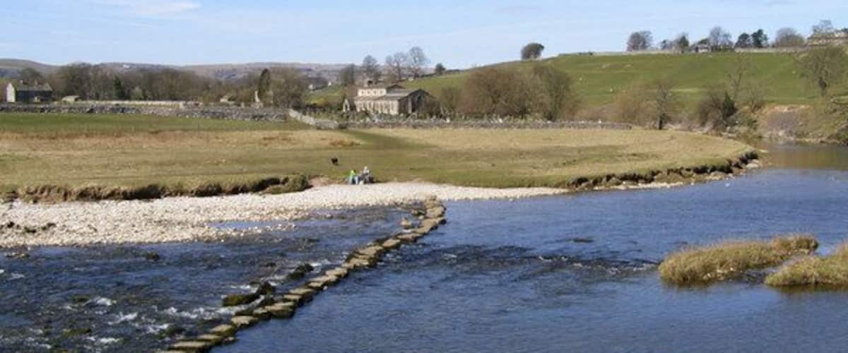 Un río con un muro de contención de piedra, riberas cubiertas de hierba y un edificio a lo lejos.