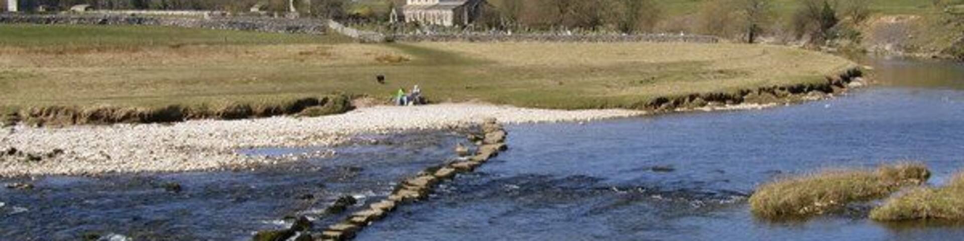 A river with a stone weir, grassy banks, and a distant building.