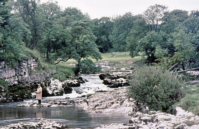 A rocky riverbank with a person standing by the water, surrounded by dense green trees.