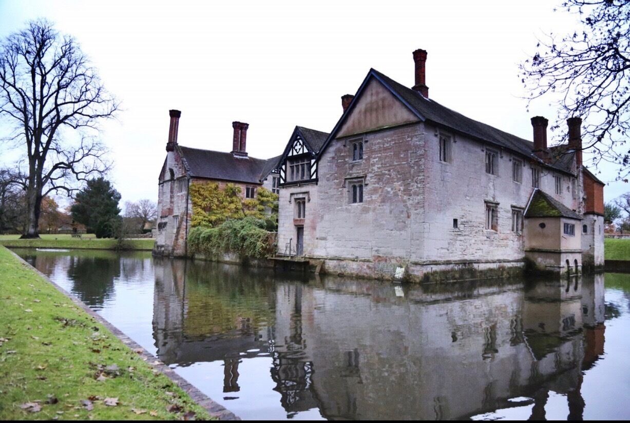 A historic stone building with a reflection in a calm water body, surrounded by trees and a grassy area.