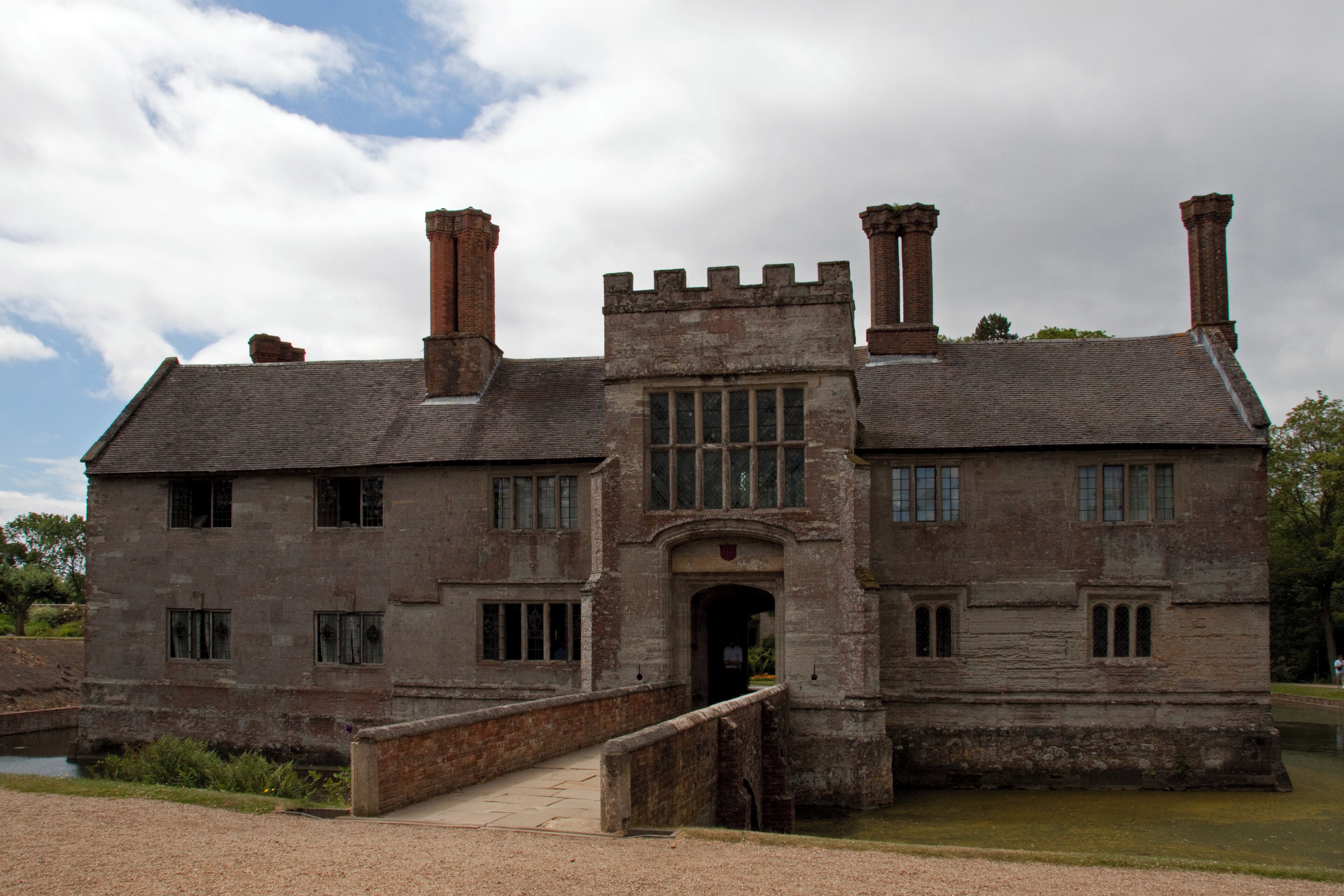 A historic stone building with a large central archway, two chimneys, and a small bridge leading to a water feature.