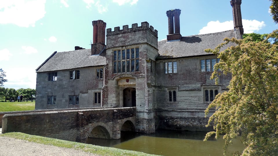 A historic stone building with a bridge over a pond, surrounded by trees.