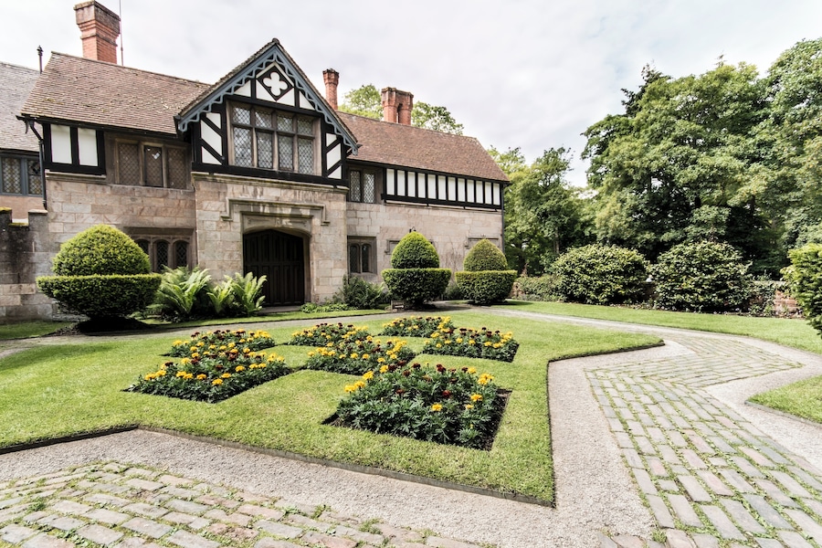 Um edifício em estilo Tudor com um jardim simétrico, com canteiros de flores e sebes bem aparadas.