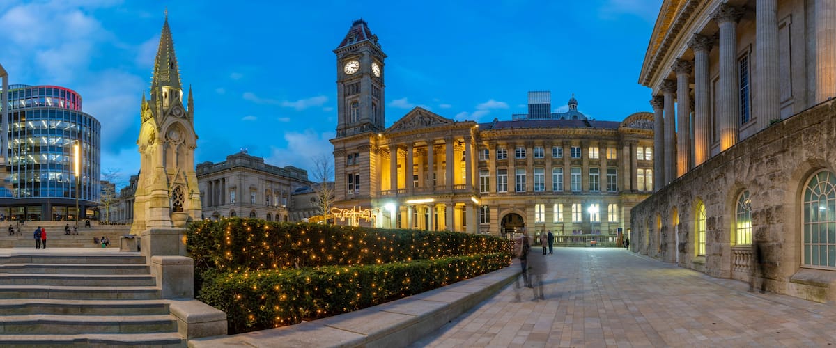 A historic square with a clock tower, a modern building, and a well-lit garden.