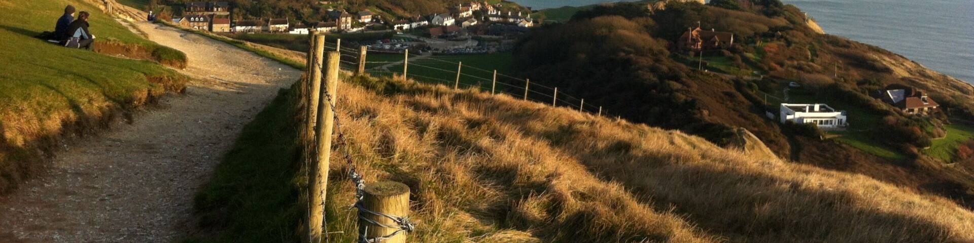 A coastal landscape with rolling hills, a sea view, and a path leading to a small settlement.