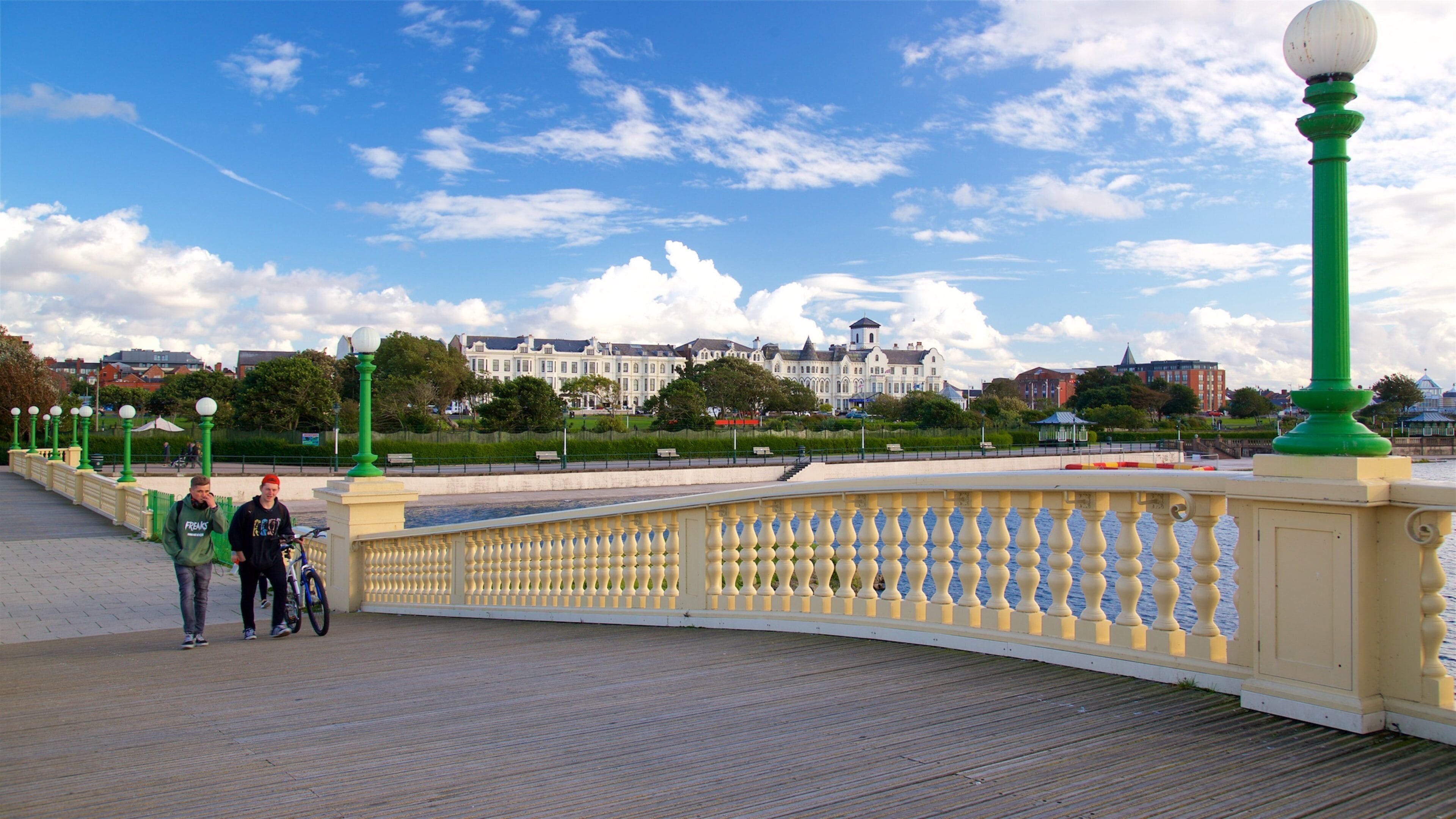 A wooden boardwalk with a green and yellow balustrade, a large white building in the background, and a clear blue sky with clouds.