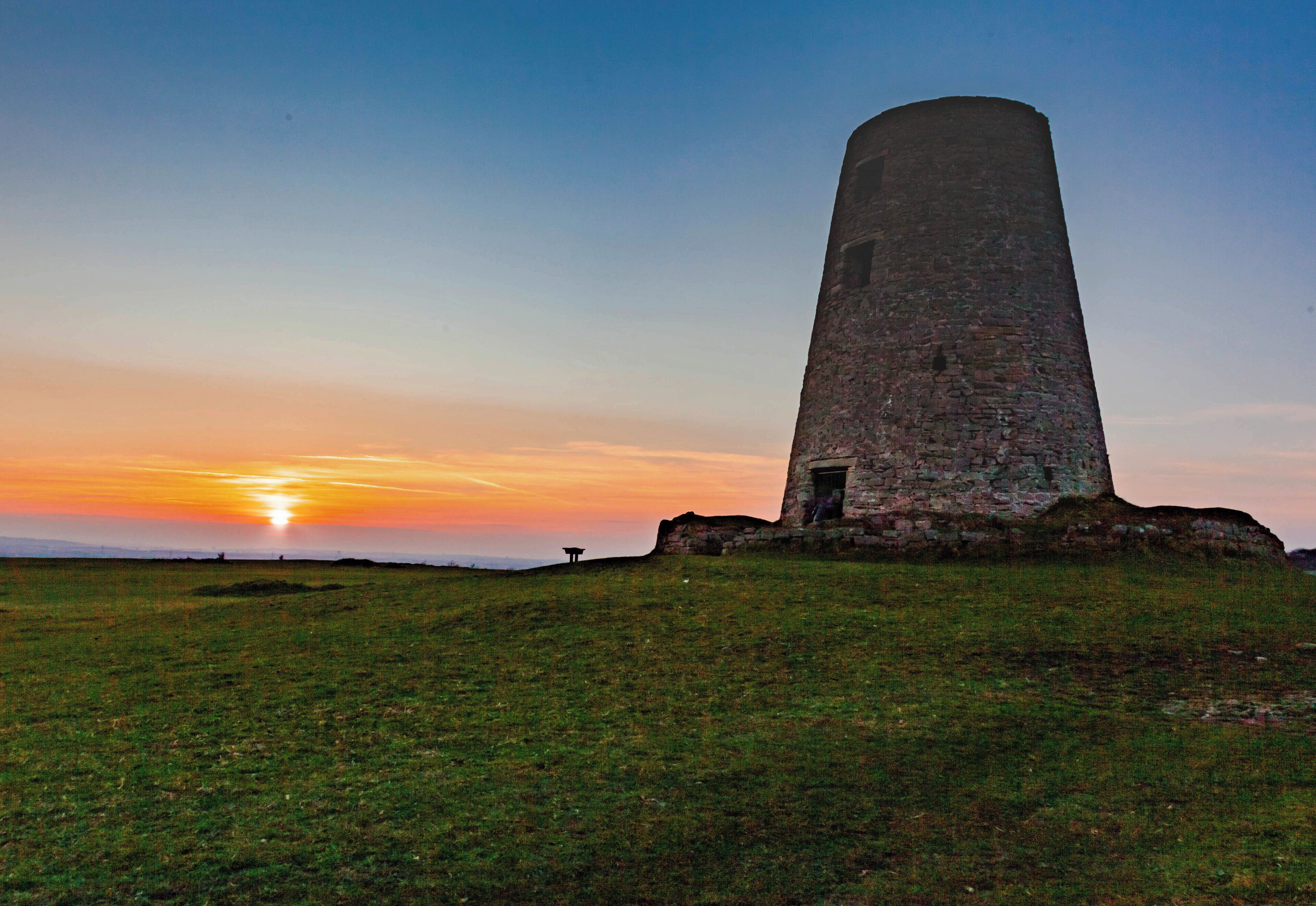A tall stone tower on a grassy hill with the sun setting in the background.