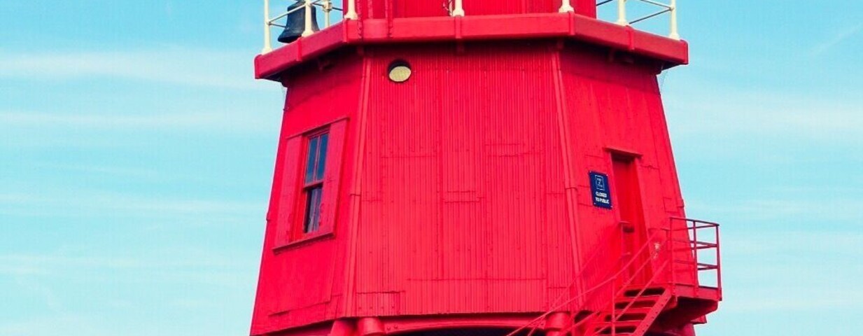Un faro rojo con veleta en la parte superior, ubicado en un muelle con vista al mar y otro faro a lo lejos.