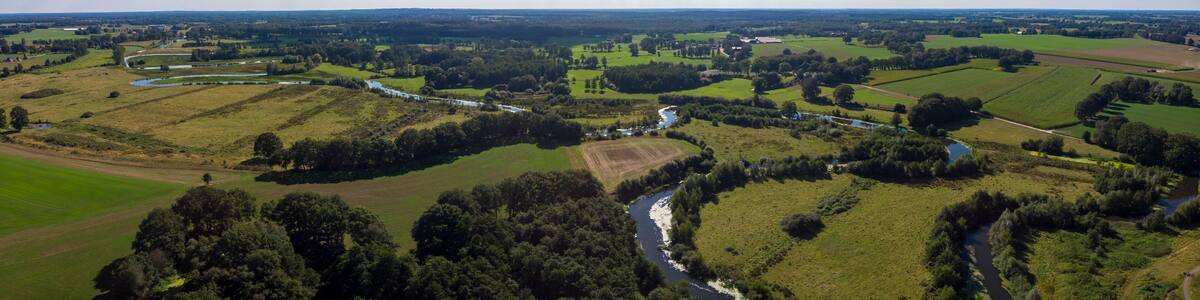 Wide aerial panorama of the river Regge meandering through the green Dutch Twente farmland landscape against a blue sky and clouds