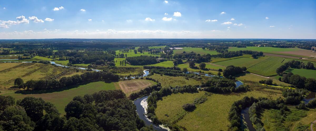 Wide aerial panorama of the river Regge meandering through the green Dutch Twente farmland landscape against a blue sky and clouds