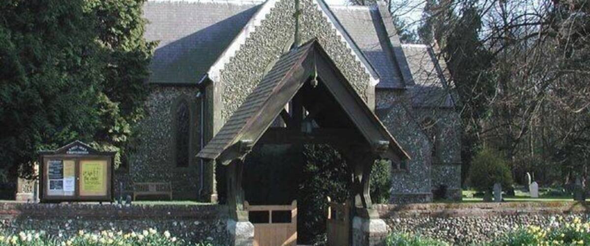 A stone building with a triangular roof and a small porch. There is a pathway leading to the entrance, and a signboard is visible to the left.