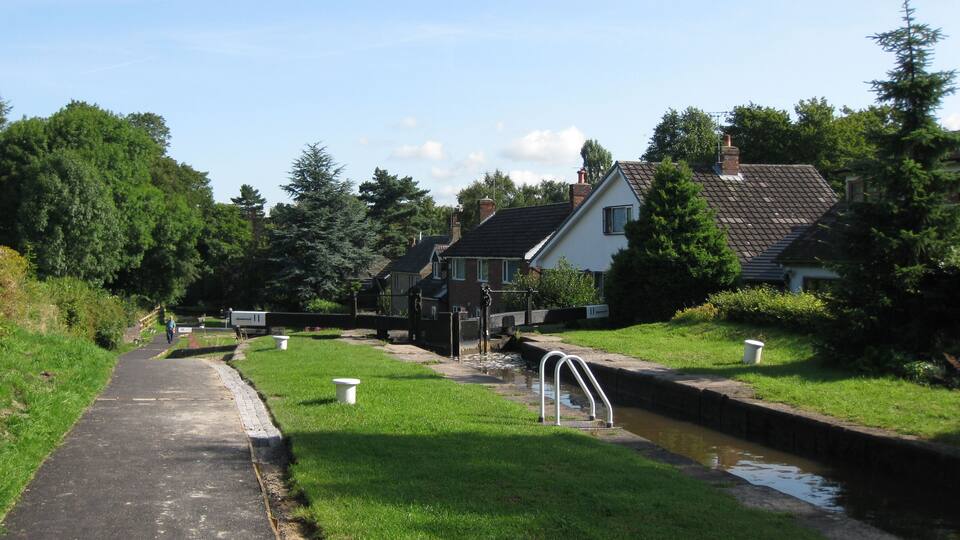 A narrow canal with a paved path alongside, surrounded by lush greenery and residential houses.