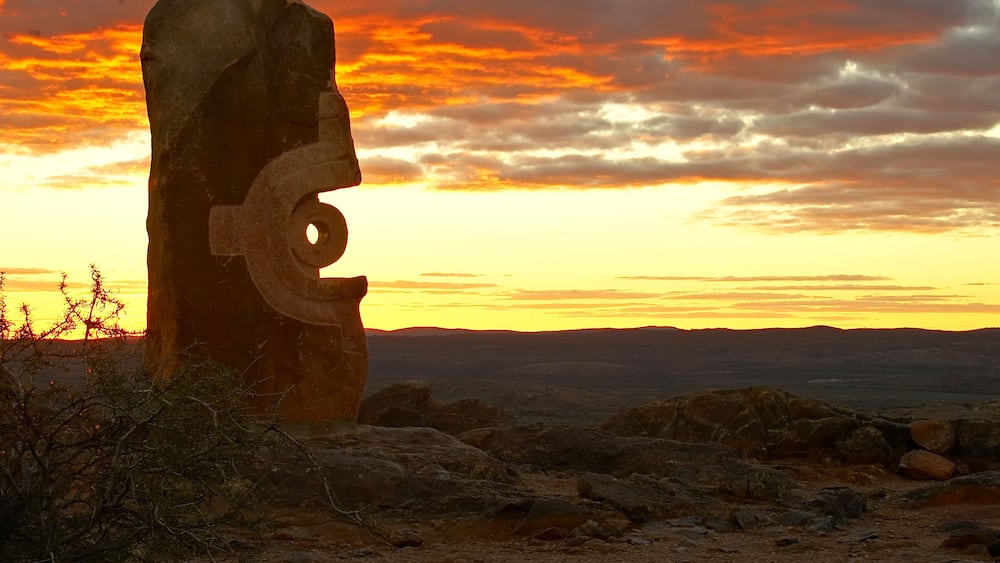 Une grande tête de pierre, dotée d’un œil et d’une bouche circulaires, se détachant sur un ciel crépusculaire parsemé de nuages.