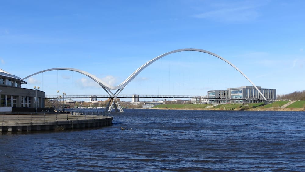 Eine moderne Brücke mit markantem Bogen über einem Fluss, mit Gebäuden und einem Steg auf der linken Seite.