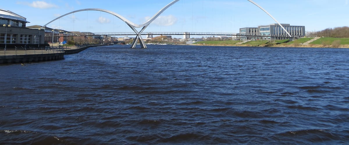 A modern bridge with a unique arch design spanning a river, surrounded by buildings and greenery.