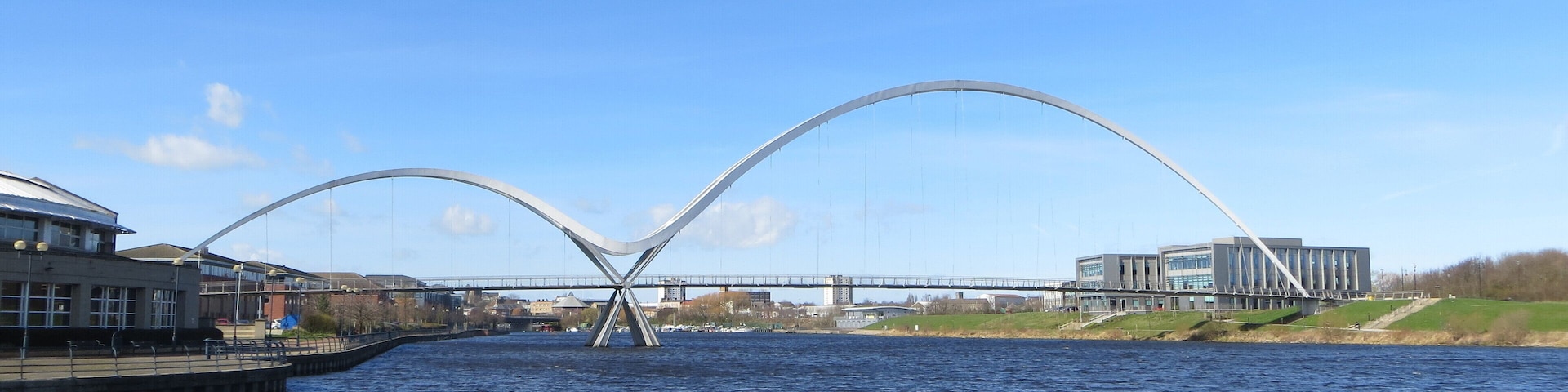 A modern bridge with a unique arch design spanning a river, surrounded by buildings and greenery.