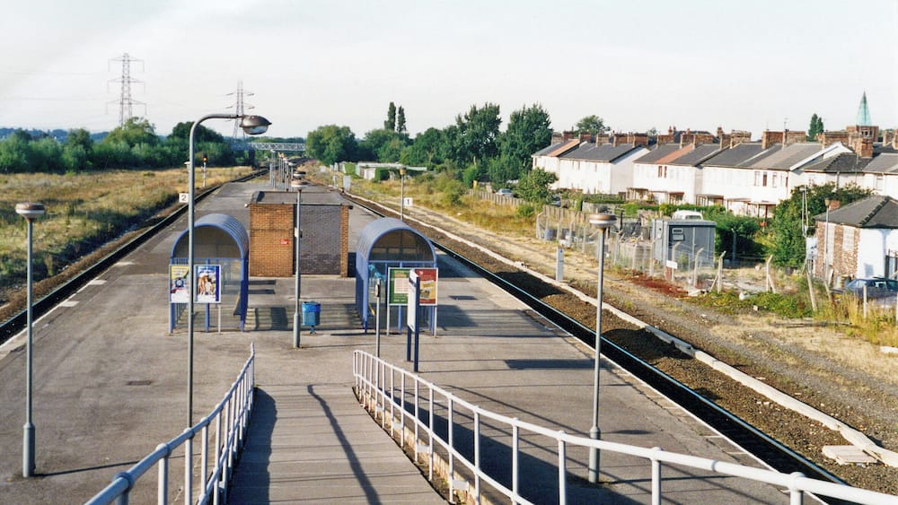 Un quai de gare avec un pont piétonnier, un poste de signalisation et des maisons d’habitation en arrière-plan.